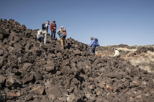 Six geologists wearing backpacks and sun protection navigate a rocky landscape while collecting geological data. The group is spread across uneven terrain under a clear blue sky, with sparse vegetation visible in the background.